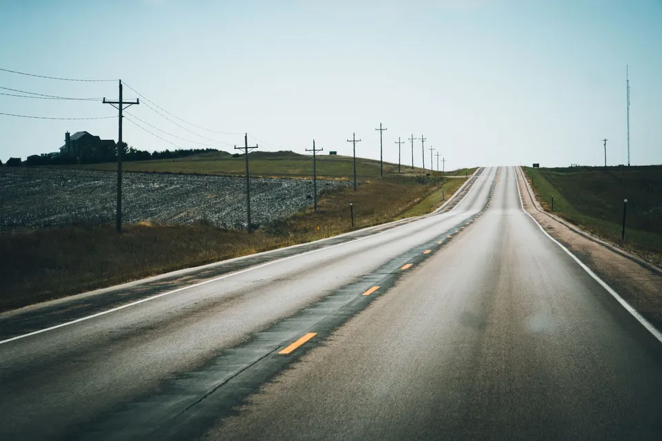 A peaceful rural highway stretching into the distance, flanked by telephone poles and open fields under a clear summer sky.