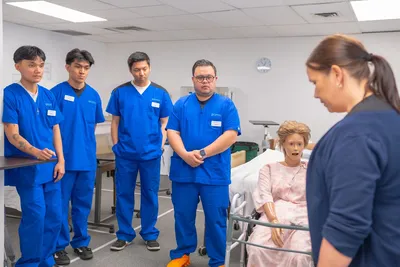 Nursing students in blue scrubs practicing patient care using a medical mannequin.