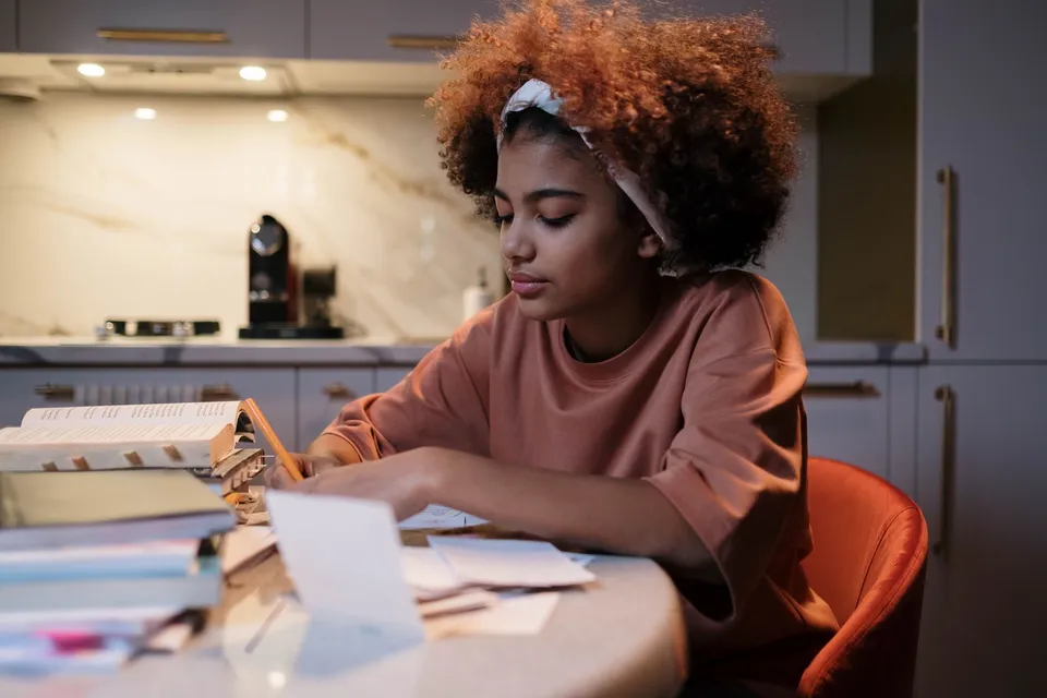 Young woman with curly hair studying at a kitchen table, surrounded by books and notes.